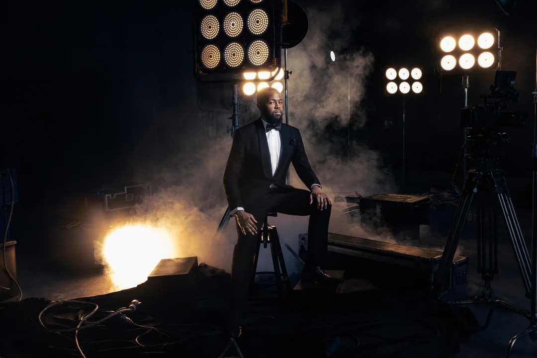 Man in a black tuxedo seated in a smoky studio among film lights